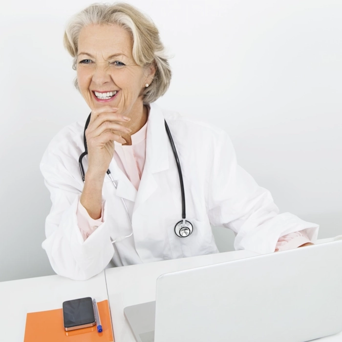 Smiling female doctor with gray hair wearing a white coat and stethoscope, sitting at a desk with a laptop